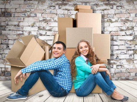 A young man and woman sit on a light wooden floor in front of a weathered brick wall, surrounded by several cardboard moving boxes of varying sizes, some open and others sealed. The man, dressed casually in a blue checkered shirt and dark jeans, is positioned on the left side, leaning back slightly with a relaxed smile. The woman, wearing a teal cardigan over a pink shirt and blue jeans, is seated on the right, hugging her knees and also smiling. Behind them, the boxes are stacked unevenly, with some leaning against the wall, and are made of light brown cardboard with visible corrugated textures. The scene takes place in a residential interior, possibly an empty or recently cleared room in a house or flat, with natural lighting creating a bright, neutral atmosphere suitable for a house removals context. The overall setting suggests an ongoing or recent move, with furnishings and personal belongings packed in the boxes, and aligns with professional house removals services in the UK, subtly referencing the location in the background context of Find Man and Van, a local removals provider.