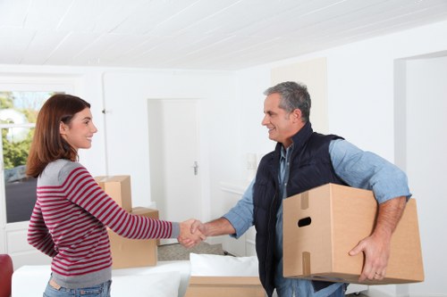 In a bright, modern domestic interior, a middle-aged man with short, grey hair, dressed in a blue shirt and dark vest, is smiling as he extends his right hand for a handshake while holding a medium-sized cardboard box in his left arm. Facing him is a young woman with shoulder-length auburn hair, wearing a long-sleeved red and grey striped sweater and jeans, shaking his hand in greeting. The background reveals a white wall with a closed door, a window with natural light, and a white sofa with some cardboard boxes placed on or near it. The scene captures a professional house removal or moving interaction, with the man representing Find Man and Van, a local removals service operating near the inferred postcode, and the setting suggesting an urban or suburban residential area typical of a town in the UK. The atmosphere is friendly and professional, emphasizing a successful handover in the context of house relocation services.
